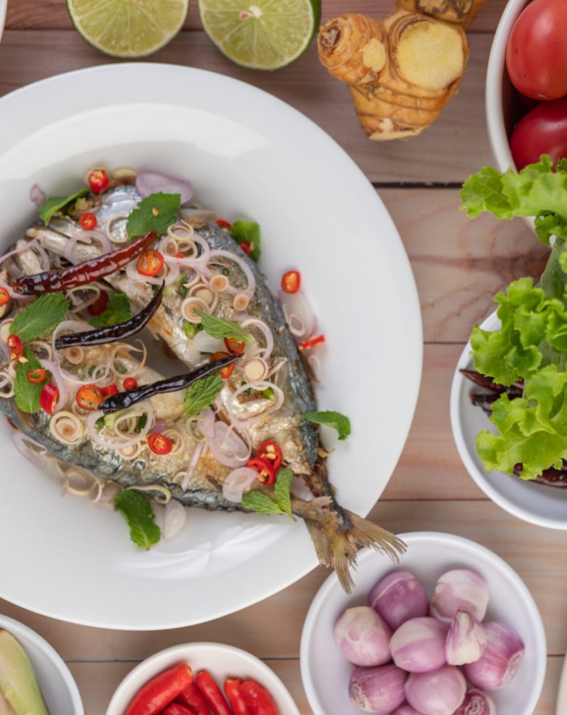 Deep-fried mackerel topped with galangal, pepper, mint, red onions in a white dish with a variety of vegetables.